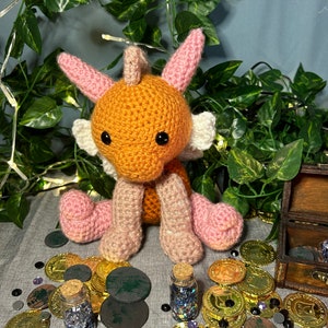 Puede incluir: Un dragón de crochet naranja y rosa con ojos negros se sienta sobre una superficie gris rodeado de monedas de oro, fichas circulares negras y verdes y pequeñas botellas de vidrio llenas de purpurina.