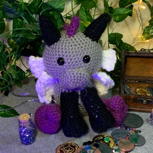 May include: A gray crocheted dragon with purple horns, black legs, and white wings. The dragon is sitting on a gray surface with gold and silver coins scattered around it.