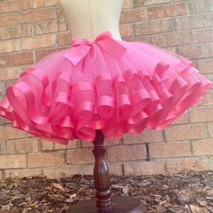 May include: A pink tulle skirt with multiple layers of pink satin ribbon. The skirt is on a wooden display stand.