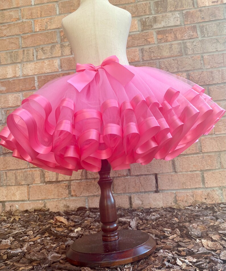 May include: A pink tulle skirt with multiple layers of pink satin ribbon. The skirt is on a wooden display stand.