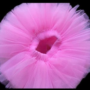 May include: A close-up of a pink tutu skirt. The skirt is made of layers of soft, sheer tulle fabric. The elastic waistband is visible at the top, and the skirt flares out in a full, circular shape. The background is black.
