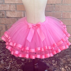 May include: A pink tulle tutu skirt with multiple layers of pink satin ribbon trim. The skirt is tied with a large pink satin bow at the back.