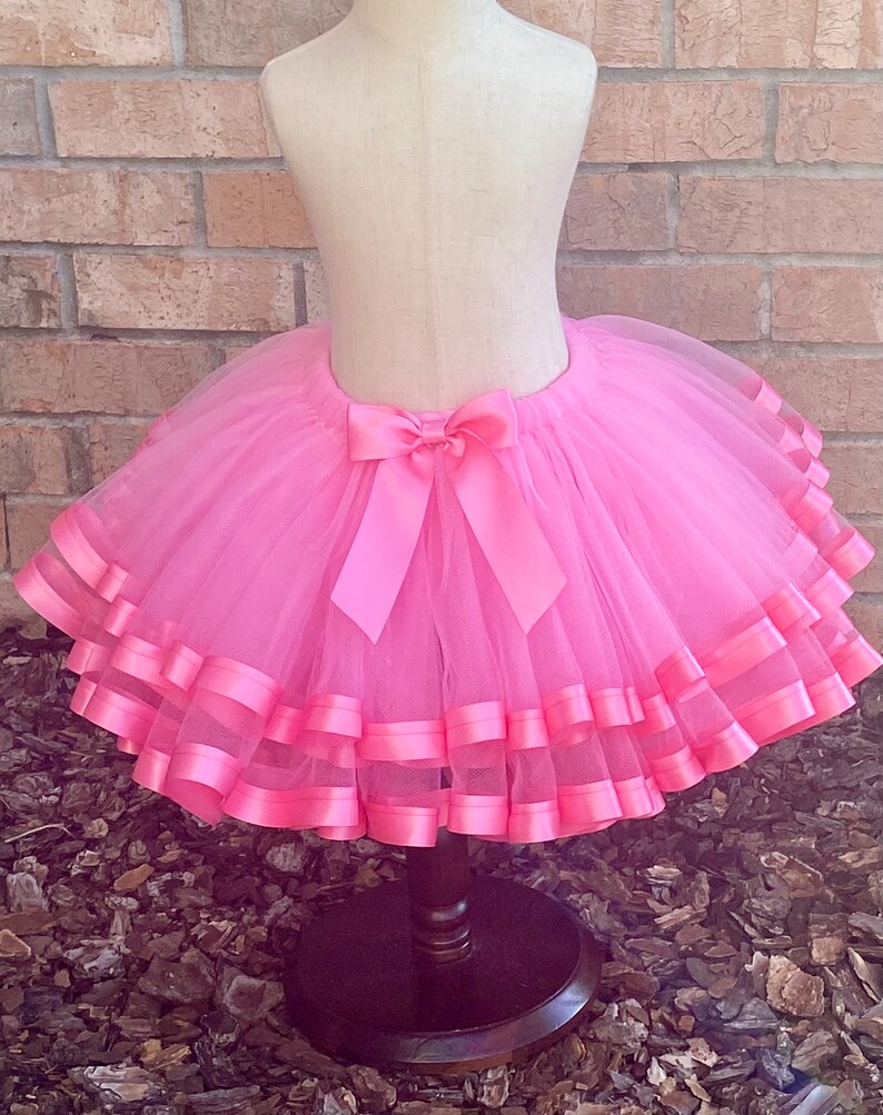 May include: A pink tulle tutu skirt with multiple layers of pink satin ribbon trim. The skirt is tied with a large pink satin bow at the back.