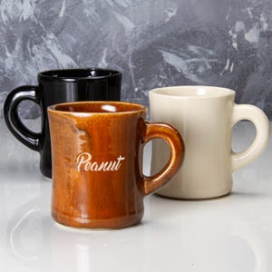 Puede incluir: Tres tazas de cerámica. Una negra, una crema y una marrón con la palabra "Peanut" en escritura blanca. Todas las tazas tienen un asa redondeada. El fondo es gris moteado.