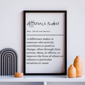 May include: A black and white printable poster with the definition of a "difference maker". The poster is framed and hanging on a white wall with decorative vases and a wooden bowl on a white shelf.
