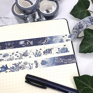 Peut inclure: Assortiment de rouleaux de ruban adhésif washi bleu et blanc sur le thème de l'océan. Le ruban présente des créatures marines comme des méduses, des hippocampes et des poissons. Le ruban est présenté sur une page de carnet à points avec un stylo noir.