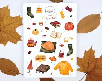 Op de afbeelding: Een stickervel met herfstillustraties. Het bevat een pompoen, een slapende kat, een kopje thee, een trui, een boek en andere herfstthema-elementen. De tekst "Cozy Autumn" is ook aanwezig.