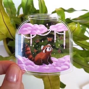 Könnte beinhalten: Ein Aufkleber in Form eines durchsichtigen Glasbehälters mit einem Roten Panda, der auf rosa Wolken sitzt. Der Behälter ist mit einer Perlenkette und Schmetterlingen verziert.
