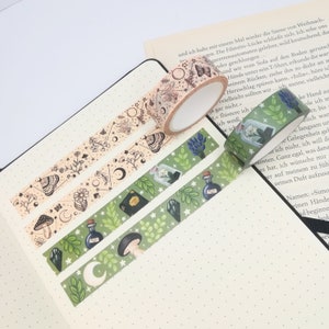 Op de afbeelding: Drie decoratieve washi-tape-rollen met hemelse en botanische ontwerpen. Eén rol heeft vlinders en sterren, een andere manen en bloemen, en de derde toont drankflessen en paddenstoelen. De tapes liggen op een gestippelde notitieboekpagina.