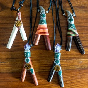 Pode incluir: Uma coleção de colares com pingentes artesanais com designs únicos. Os pingentes apresentam vários materiais, incluindo madeira, pedra e metal, com padrões intrincados e detalhes coloridos. Cada colar pende de um cordão preto, mostrando os pingentes.