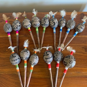 May include: A selection of handcrafted percussion instruments. Each instrument has a dark, patterned gourd head on a wooden handle. The gourds are decorated with detailed designs and topped with feathers. The handles are wrapped with colourful threads.