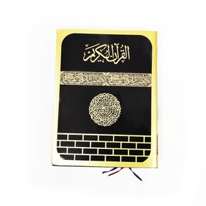 May include: A black and gold Quran with Arabic calligraphy. The cover features gold accents and a decorative design. The book has a rectangular shape with a gold border and a white background. The title "Al-Quran Al-Kareem" is visible on the cover.