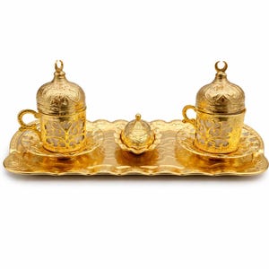 Puede incluir: Un juego de café turco dorado, que incluye dos tazas ornamentadas con tapas, platillos y un plato pequeño, todo sobre una bandeja a juego. El juego presenta intrincados patrones en relieve y un acabado brillante y reflectante, ideal para servir café.