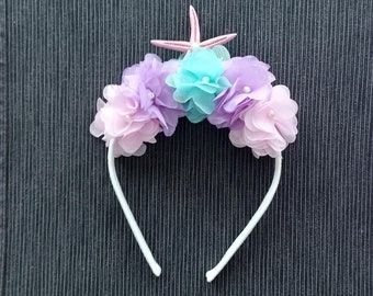 Puede incluir: Una diadema blanca adornada con un grupo de flores de tela superpuestas en tonos morado claro, lavanda y turquesa. Cada flor presenta una pequeña cuenta blanca en el centro. Un adorno de estrella de mar rosa pálido se sitúa arriba.
