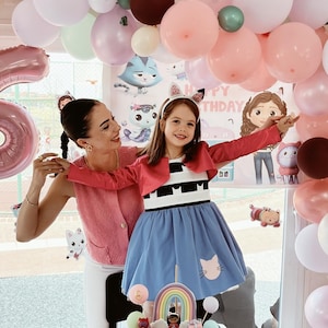 May include: A young girl in a blue dress with a cat design and a red jacket poses with a woman in front of a birthday party backdrop. The party features balloons in various colors and a decorated cake with a rainbow.