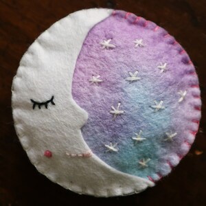 Puede incluir: Una luna creciente de fieltro blanco con un ojo cerrado y un rubor rosado, bordada con estrellas blancas sobre un fondo degradado morado y azul.