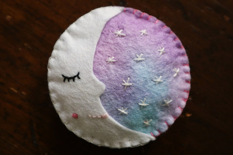 Puede incluir: Una luna creciente de fieltro blanco con un ojo cerrado y un rubor rosado, bordada con estrellas blancas sobre un fondo degradado morado y azul.