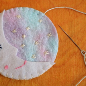 Puede incluir: Una luna creciente de fieltro blanco con un fondo degradado rosa y azul y estrellas bordadas blancas. La luna tiene un ojo bordado negro y una boca bordada rosa. La luna se est&aacute; cosiendo con hilo blanco.