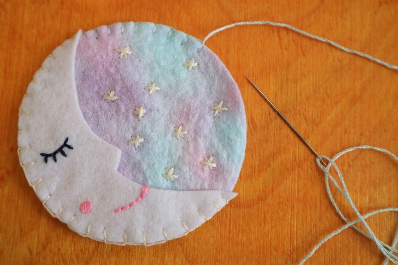 Puede incluir: Una luna creciente de fieltro blanco con un fondo degradado rosa y azul y estrellas bordadas blancas. La luna tiene un ojo bordado negro y una boca bordada rosa. La luna se est&aacute; cosiendo con hilo blanco.