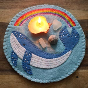 May include: A blue felt whale with white stitching and white dots, a rainbow, a lit candle in a seashell, and sea glass on a blue felt background.
