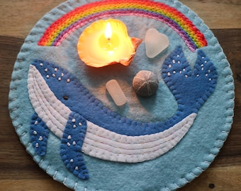 Puede incluir: Una ballena azul de fieltro con costuras blancas y puntos blancos, un arcoíris, una vela encendida en una concha de mar y vidrio de mar sobre un fondo azul de fieltro.