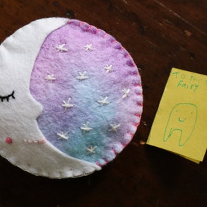Puede incluir: Una luna de fieltro blanco con un fondo degradado rosa, morado y azul y estrellas bordadas blancas. La luna tiene un ojo bordado negro y una boca bordada rosa. Una nota amarilla con el texto "To the Tooth Fairy" y un dibujo verde de un diente con una cara sonriente.