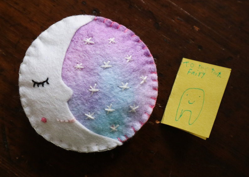 Puede incluir: Una luna de fieltro blanco con un fondo degradado rosa, morado y azul y estrellas bordadas blancas. La luna tiene un ojo bordado negro y una boca bordada rosa. Una nota amarilla con el texto "To the Tooth Fairy" y un dibujo verde de un diente con una cara sonriente.