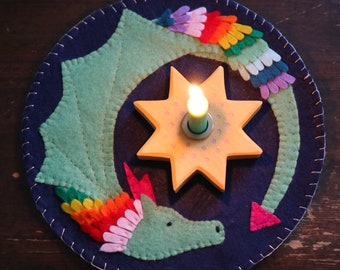 Puede incluir: Un portavelas de fieltro con un diseño de dragón verde. El dragón tiene una melena y una cola arcoíris. El portavelas está decorado con una estrella amarilla con una vela en el centro.