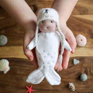 May include: A white plush mermaid doll with a white crocheted hat and long white braids. The doll is wearing a white dress and has a painted face with pink cheeks. The doll is being held by a person's hands.
