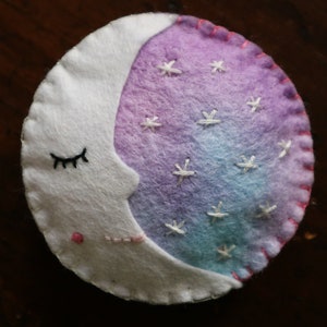 Puede incluir: Una luna creciente de fieltro blanco con una cara dormida y costuras rosas. La luna est&aacute; medio llena de fieltro degradado morado y azul con estrellas bordadas blancas.