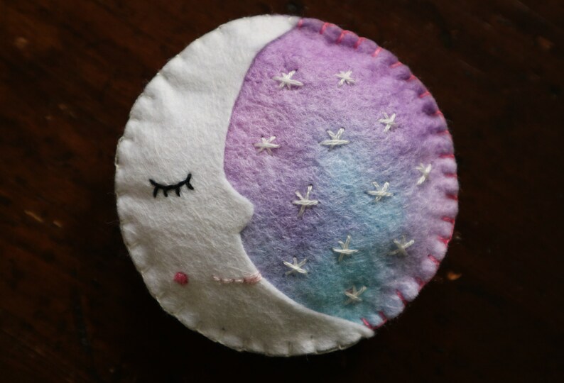Puede incluir: Una luna creciente de fieltro blanco con una cara dormida y costuras rosas. La luna est&aacute; medio llena de fieltro degradado morado y azul con estrellas bordadas blancas.