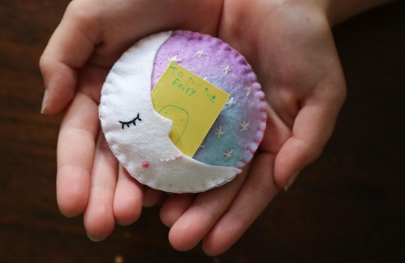 Puede incluir: Una luna creciente de fieltro blanco con pesta&ntilde;as bordadas sostiene una nota amarilla que dice "To the Tooth Fairy". La luna est&aacute; decorada con fieltro rosa y morado y estrellas bordadas blancas.