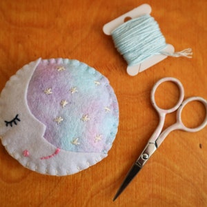 Puede incluir: Una luna de fieltro con forma de media luna blanca y estrellas y pesta&ntilde;as bordadas. La luna es azul claro y morada con un contorno blanco. Hay una bobina de hilo azul claro y un par de tijeras plateadas sobre una superficie de madera.