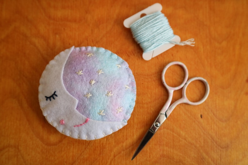 Puede incluir: Una luna de fieltro con forma de media luna blanca y estrellas y pesta&ntilde;as bordadas. La luna es azul claro y morada con un contorno blanco. Hay una bobina de hilo azul claro y un par de tijeras plateadas sobre una superficie de madera.