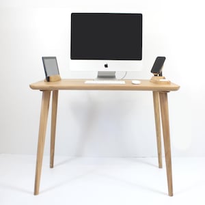 Può includere: Una scrivania in legno chiaro con computer, tastiera e mouse. Un tablet e uno smartphone poggiano su supporti in legno. La scrivania ha quattro gambe angolate e un bordo arrotondato.