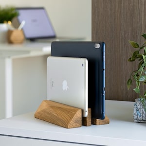 Peut inclure: Support en bois pour tablette, tenant une tablette argentée et une tablette bleu foncé. Le support est en bois brun clair et présente un design géométrique. Les tablettes sont sur une surface blanche, avec un bureau et des plantes en arrière-plan.