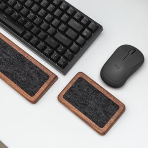 May include: A black keyboard, mouse, and two wooden wrist rests with gray felt inserts are arranged on a white surface. The wrist rests have a rectangular shape and a dark wood frame.