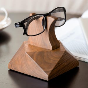 Könnte beinhalten: Ein Brillenständer aus Holz mit geometrischer Basis und geschwungener Oberseite, der eine Brille mit schwarzem Rahmen hält. Der Ständer ist aus warmtonigem Holz gefertigt und steht auf einer dunklen Oberfläche.