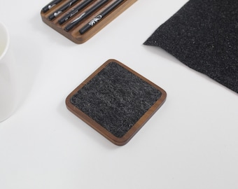 May include: A square coaster with a dark gray felt insert and a wooden frame. A wooden chopstick holder with chopsticks and a piece of dark gray fabric are also visible. The items are on a white surface.