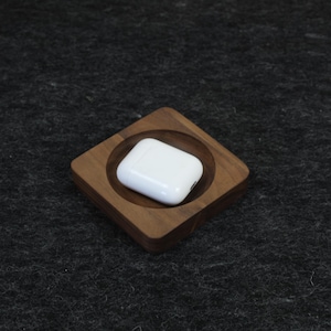 Może przedstawiać: Białe bezprzewodowe etui ładujące do słuchawek leży w kwadratowej drewnianej miseczce z zaokrąglonym środkiem.