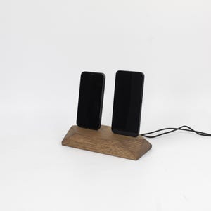 May include: A wooden phone stand holding two black smartphones. The stand is made of light brown wood and has a geometric design. A black charging cable is connected to the stand. The background is white.