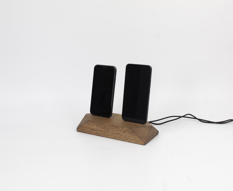 May include: A wooden phone stand holding two black smartphones. The stand is made of light brown wood and has a geometric design. A black charging cable is connected to the stand. The background is white.