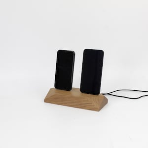 May include: A wooden phone charging station holding two black smartphones. The station is made of light brown wood with a geometric design. A black charging cable extends from the base.