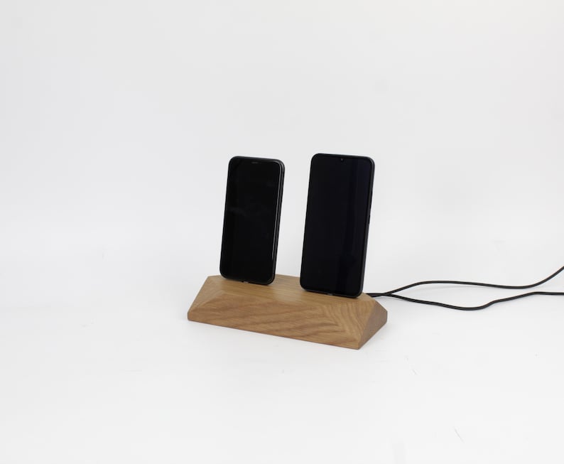 May include: A wooden phone charging station holding two black smartphones. The station is made of light brown wood with a geometric design. A black charging cable extends from the base.