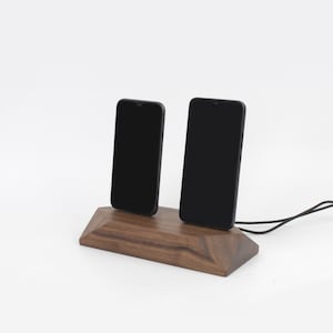 May include: A wooden phone charging station with two black smartphones. The station is made of dark brown wood and has a geometric design. A black charging cable extends from the base.