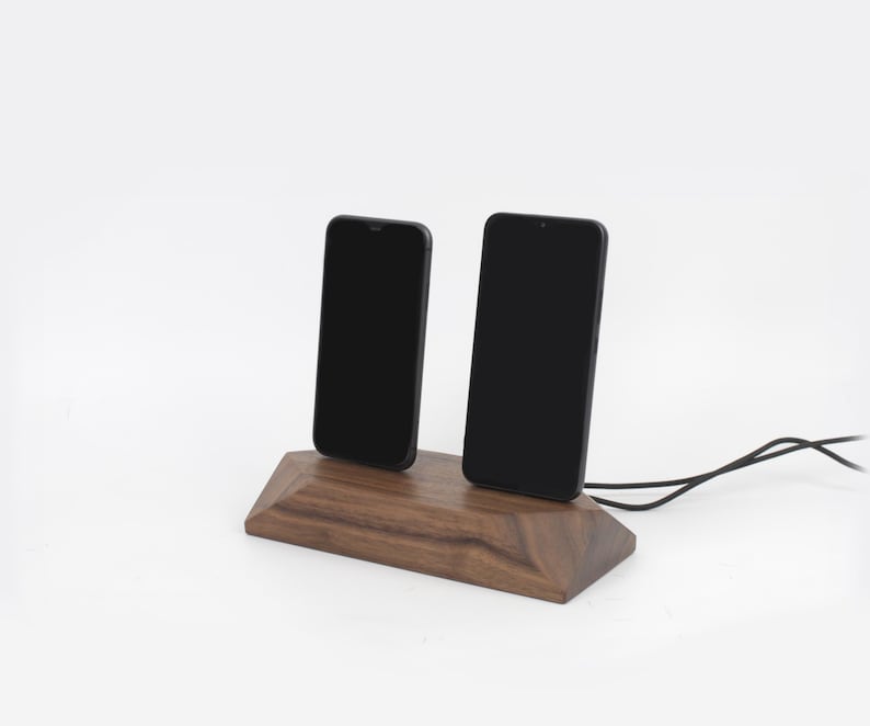 May include: A wooden phone charging station with two black smartphones. The station is made of dark brown wood and has a geometric design. A black charging cable extends from the base.