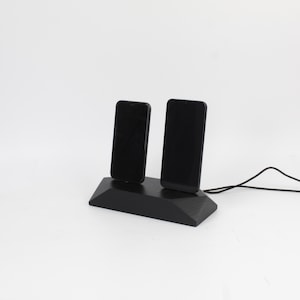 May include: A black wooden phone charging station holding two smartphones. The phones are upright and connected to a black cable. The charging station has a geometric design, set against a white backdrop.