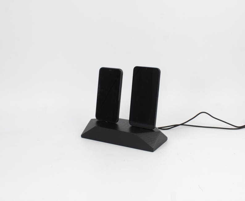 May include: A black wooden phone charging station holding two smartphones. The phones are upright and connected to a black cable. The charging station has a geometric design, set against a white backdrop.