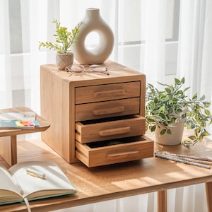 May include: A wooden desk organiser with three drawers, one open. The natural wood finish complements the other items on the desk, including a white vase with a circular design, a small plant, and a notebook with a pen. The desk is made of wood.