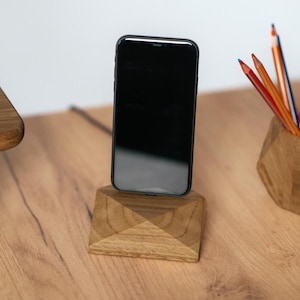 Könnte beinhalten: Ein Holz-Telefonständer mit geometrischem Design, der ein schwarzes Smartphone hält. Der Ständer steht auf einer hellbraunen Holzoberfläche.